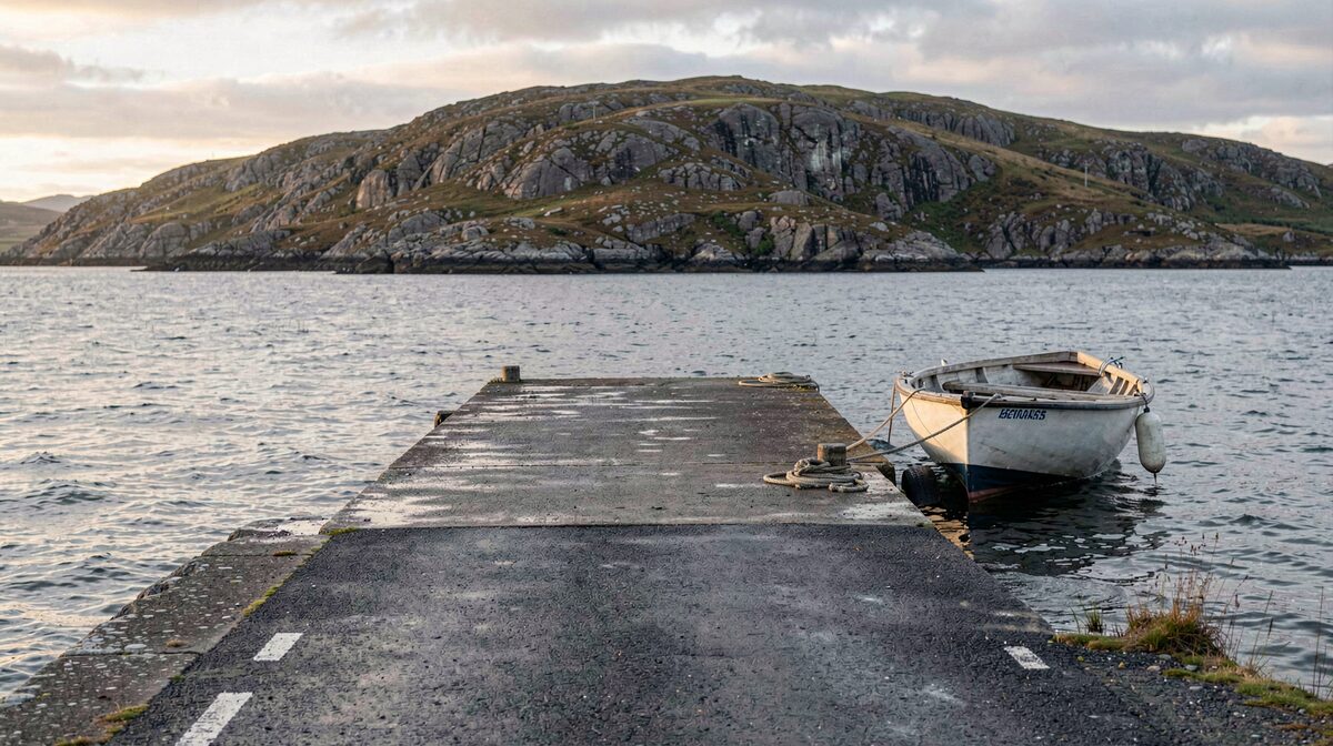 A single-track road ending at a concrete slipway on a Hebridean loch, a small open boat moored at the edge, low evening light on wet tarmac and grey water, wide framing with a headland behind.
